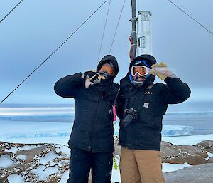 Two men with their faces covered by large goggles and hoods are holding up sausages to their mouths. Behind them is the steel mast of a weather station, and the background shows a large expanse of snow and ice.