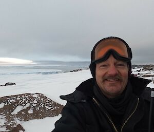 A man with goggles on top of his head is smiling at the camera. In the background is a rocky, snow covered hill, dropping away gradually to the face of a large glacier. The sky is completely overcast.