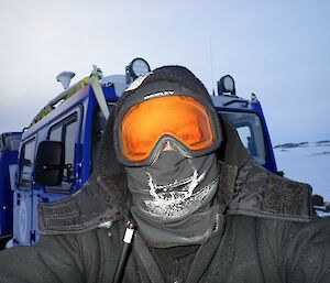 A man with his face completely covered by goggles and a face warmer is taking a selfie. In the background is a Hagglund tracked vehicle and snow covered ground. The sky is completely overcast.