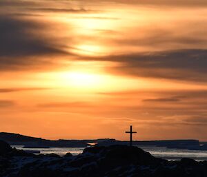 A steel memorial cross sits atop a rocky snow covered hill. In the background the sun is low in the sky and being filtered by thin cloud to create an orange hue.