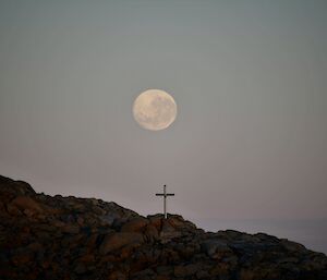 A steel memorial cross sits midway up a rocky hill (Reeves Hill) with a full moon directly above it.