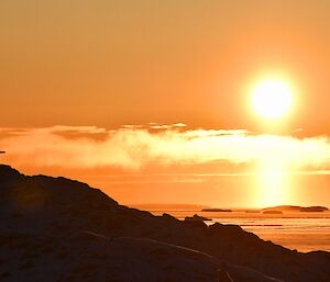 A steel memorial cross sits to the far left of the photo, near the top of a rocky hill. In the background the sun is low on the horizon, and painting the sky orange over the iceberg strewn water.