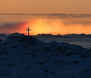 A steel memorial cross sits atop a rocky snow covered hill. In the background a rising fog off the water is being illuminated by sunlight.