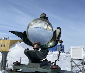 A Campbell-Stokes sunshine recorder with glass sphere is in centre frame. A blue building and a yellow building are in the background and that background image is visible refracted through the glass sphere.