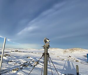 A Campbell-Stokes sunshine recorder is atop a metal pole steadied by four guy wires. In the background is a snow covered landscape and the clouds stretch across the sky.