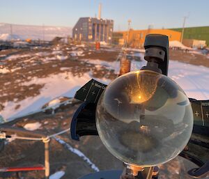 A Campbell-Stokes sunshine recorder with glass sphere is in centre frame. A reflection of a person holding a phone is visible in the sphere. There is a yellow, a blue, and a green building, and snow covered rocky land in the background.