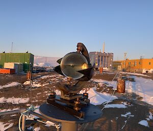 A Campbell-Stokes sunshine recorder with glass sphere is in centre frame. A reflection of the sunset is visible in the sphere. There is a yellow, a blue, and a green building, and snow covered rocky land in the background.