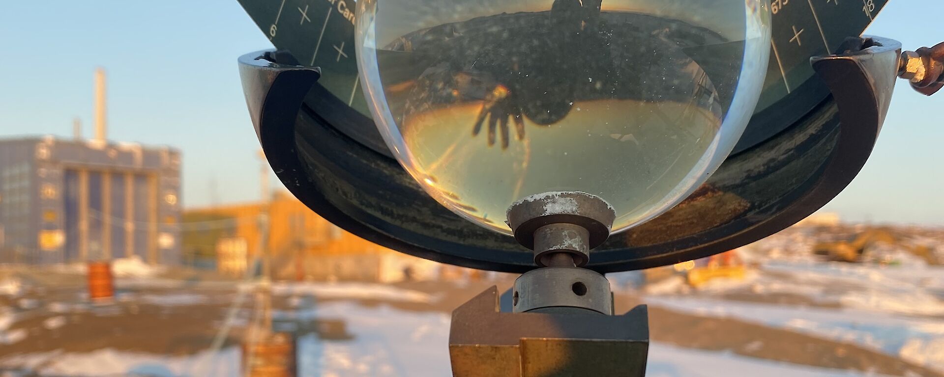 A Campbell-Stokes sunshine recorder glass sphere shows the reflection and refracted image of a person waving and a sunset. There is a blue building and snow covered rocky land in the background.