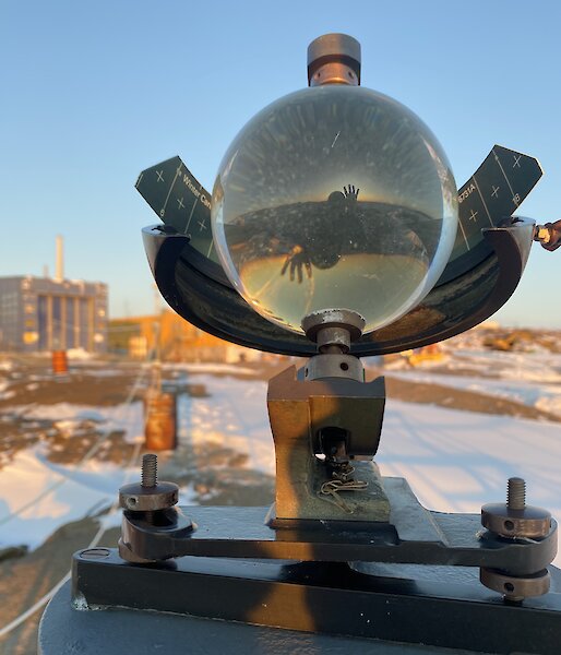 A Campbell-Stokes sunshine recorder glass sphere shows the reflection and refracted image of a person waving and a sunset. There is a blue building and snow covered rocky land in the background.