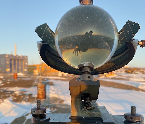 A Campbell-Stokes sunshine recorder glass sphere shows the reflection and refracted image of a person waving and a sunset. There is a blue building and snow covered rocky land in the background.