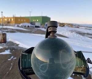 A Campbell-Stokes sunshine recorder with glass sphere is in centre frame. There is a yellow and a green building and snow covered rocky land in the background.