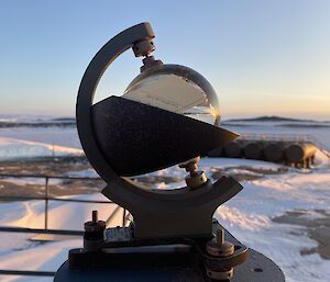 A Campbell-Stokes sunshine recorder with glass sphere is in centre frame. There are a number of large fuel tanks, an ice covered harbour, and snow covered rocky land in the background.
