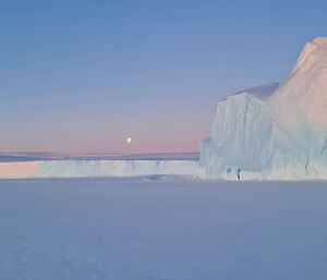 A large iceberg frozen into the sea-ice is to the right of frame. Ice cliffs and an ice plateau is in the distance and the moon is visible in the sky. Pastel blues and pinks are across the scenery.