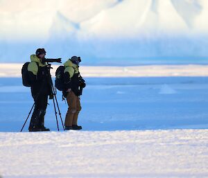 Two men are standing next to a large camera with telephoto lens on a tripod. In the distance, an iceberg rises from the sea-ice.
