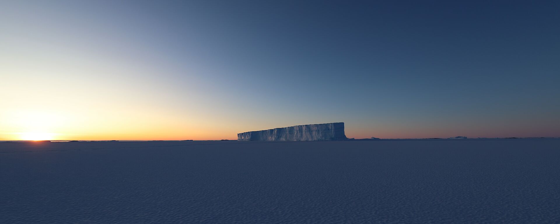 The sun is setting in the distance over the sea-ice. A large iceberg, frozen into the sea-ice, is in the centre of frame.