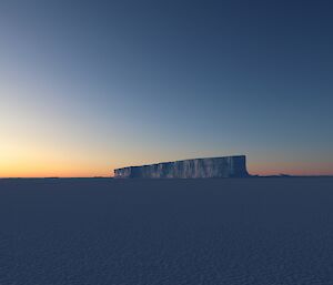 The sun is setting in the distance over the sea-ice. A large iceberg, frozen into the sea-ice, is in the centre of frame.