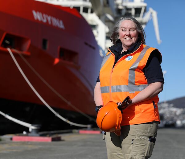 A woman in an orange vest holding an orange hard hat standing in front of the bow of a red ship with the name Nuyina