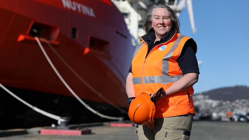 A woman in an orange vest holding an orange hard hat standing in front of the bow of a red ship with the name Nuyina