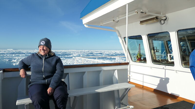 A woman in a beanie and blue fleece jacket sits outside the bridge of a ship on a sunny day on on ice-covered ocean