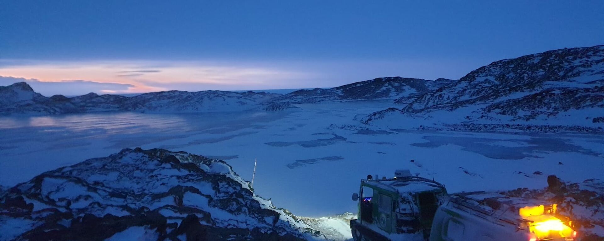 A green snow-covered vehicle sits atop a rocky landscape just in front of a snow covered frozen lake.