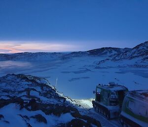 A green snow-covered vehicle sits atop a rocky landscape just in front of a snow covered frozen lake.