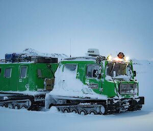 A green snow vehicle with front and rear cabins sits in a snowy landscape.