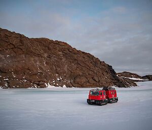 A red snow vehicle sits on the frozen fjord ice with a large rocky hill behind it.