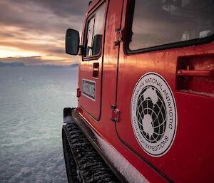 Looking down the side of a red snow vehicle (with an Australian Antarctic Division sticker on the side door) towards the snow-covered sea-ice and setting sun.