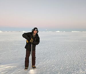 A smiling man stands on the sea-ice while drilling a hole in the ice.
