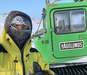 A man wearing a buff, beanie and several layers of clothing covered in ice crystals stands next to the front door of a green Hägglunds.