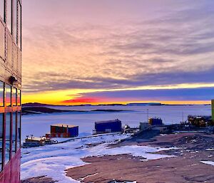 A red and orange sunset is visible on the horizon against clouds stretching across the frame. There is a large red building with reflective windows to the left of frame and colourful buildings in the middle ground on a rocky, snow covered landscape.