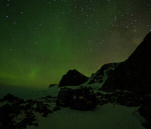 A green aurora is visible in thee night sky. A blue Hägglunds vehicle is in the centre of frame surrounded by a snow and ice covered rocky landscape.