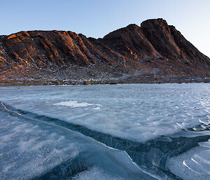 A frozen lake is seen with rippled surface and frozen air bubbles beneath. In the background is a rocky ridge line.