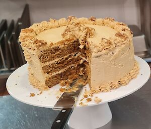 A large, decorated, coffee cake is sitting on a white cake stand. There is a knife resting on the stand.