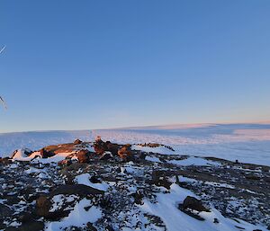 A large wind turbine is visible to the left of frame rising from a rocky, snow covered landscape. There is an ice plateau rising into the distance in the background.