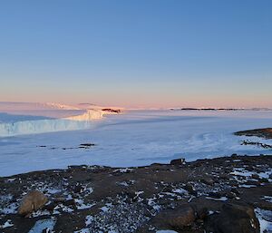 Sea ice stretches into the distance from a rocky hillside. To the left of frame, ice cliffs and an ice plateau rise. There are ice bergs and islands visible far in the distance.