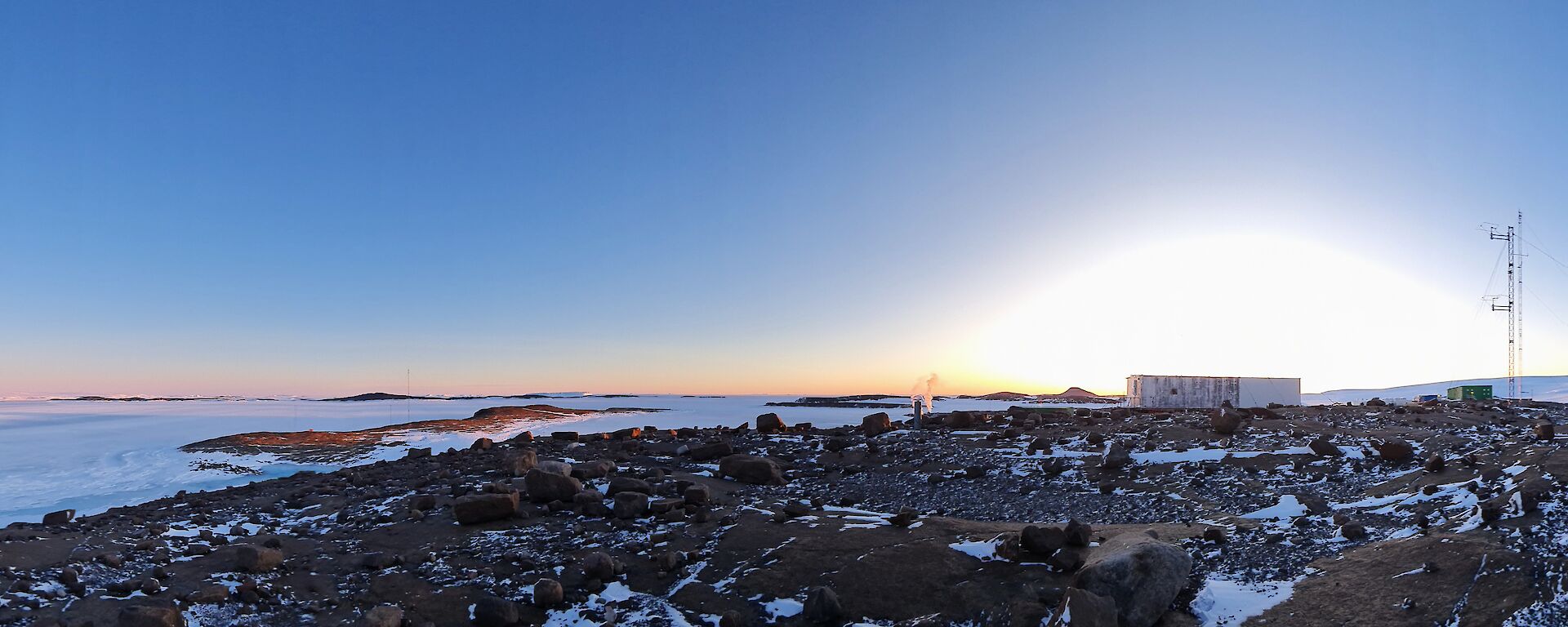 A panoramic photograph of a rocky landscape. There is an old building silhouetted by the glow of sunrise in the centre of frame and a large wind turbine to the right of frame