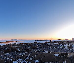 A panoramic photograph of a rocky landscape. There is an old building silhouetted by the glow of sunrise in the centre of frame and a large wind turbine to the right of frame