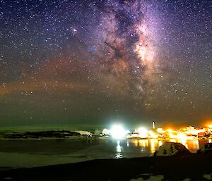 A brightly lit station is on a rocky landscape under a night sky featuring a very bright Milky Way.