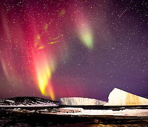 A bright red, yellow, and green aurora is visible in the night sky above two large ice bergs. There is a rocky island visible to the left of frame