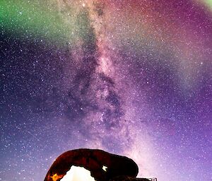 A bright green and purple aurora is visible in the night sky overlaying the Milky Way. In the foreground is a large snow covered rock.