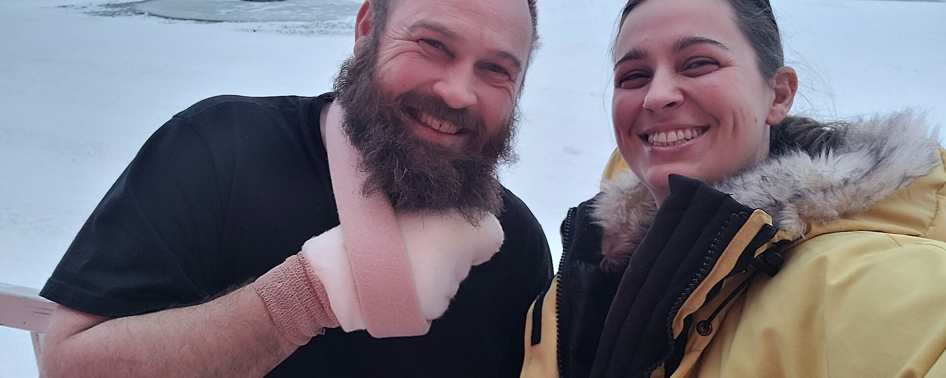 A man with his arm in a sling and hand bandaged, and a woman in a yellow winter jacket are smiling at the camera. In the background is a snow covered ground and the sun is visible low on the horizon.