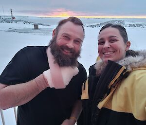 A man with his arm in a sling and hand bandaged, and a woman in a yellow winter jacket are smiling at the camera. In the background is a snow covered ground and the sun is visible low on the horizon.