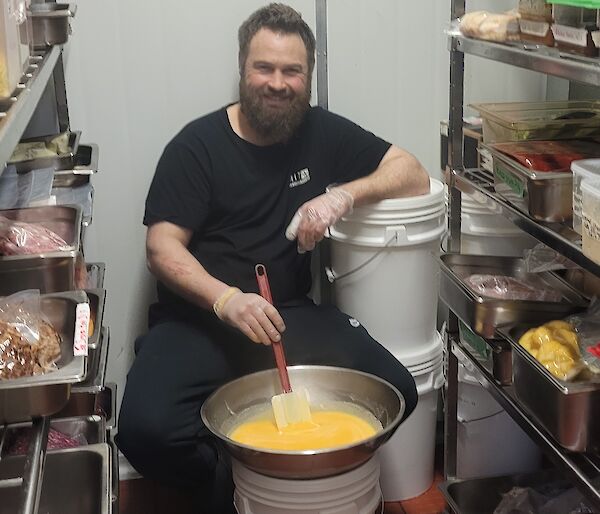 A man is sitting in between two racks of steel food trays, inside a large kitchen cool room. He is stirring a big bowl of eggs and smiling at the camera. His left index finger is bandaged.