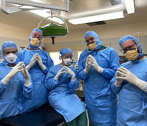 Five people in blue medical scrubs and face masks are standing in a line facing the camera with their hands held together in front of them.