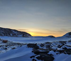 An red/orange van on sleds is parked on a rocky shore. In the distance are more rocky islands and the sea is frozen ice.