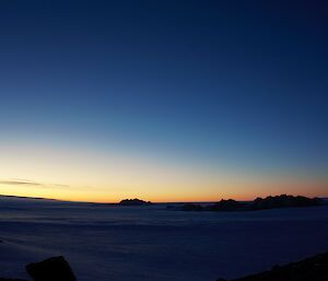 On the horizon the glow of sunrise can be seen against a dark blue sky. An ice plateeau stretchs into the distance with a rocky mountain range visible.