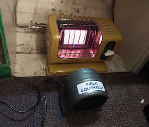 An electric heater is on the floor in front of a gas powered heater.