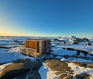 An old rectangular hut is tied down to a rocky island. In the background is a red half-dome hut and two Hägglunds vehicles.