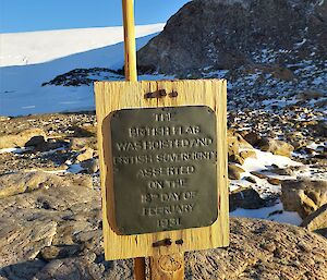 A metal plaque is mounted on a wooden board standing on a rocky landscape. There is a bamboo cane just behind the board.